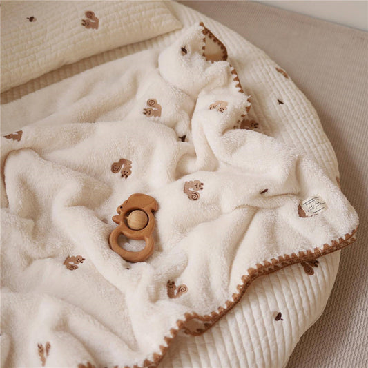 A beige baby blanket with a soft coral fleece texture, embroidered with a pattern of little bears, and a wooden teether lying on the blanket.